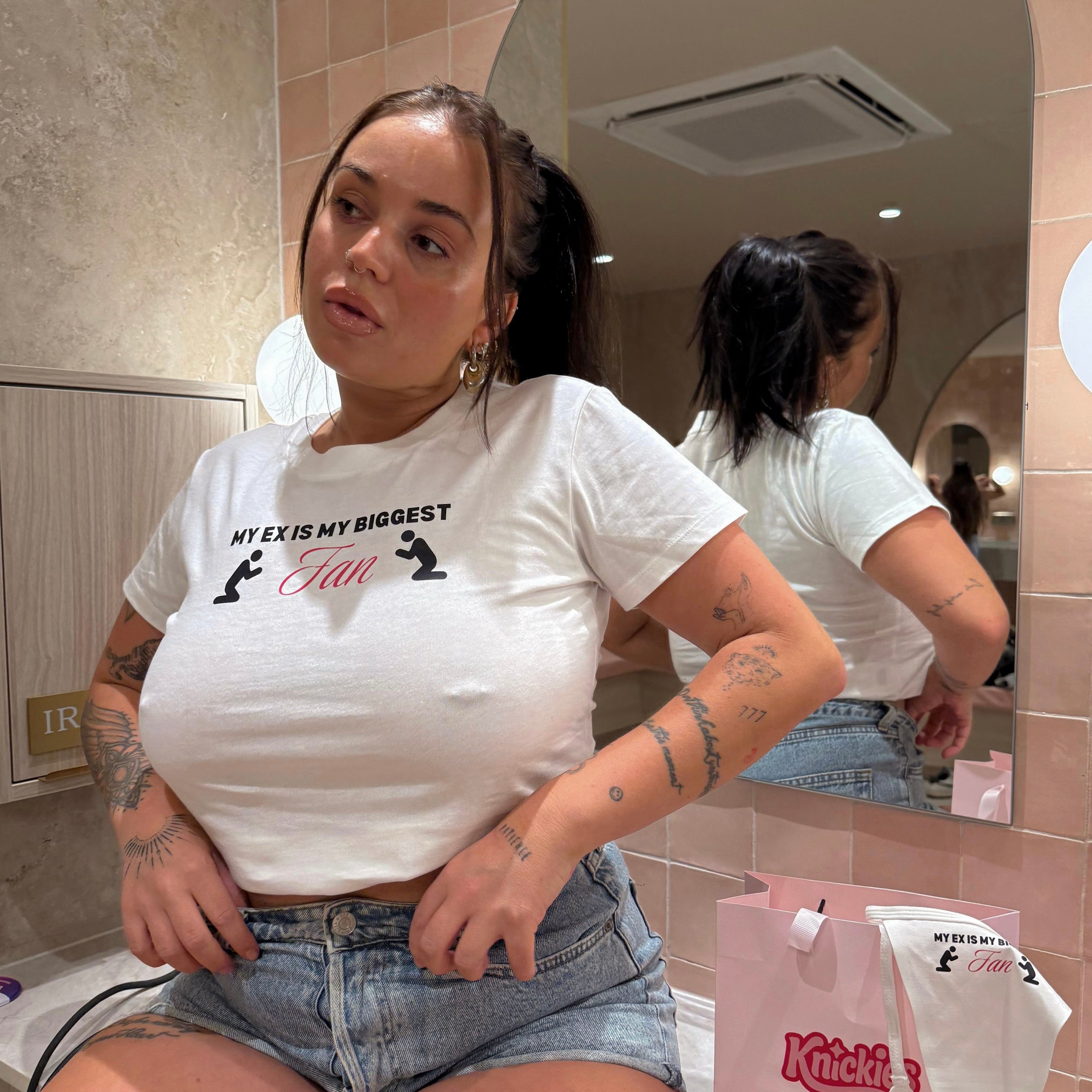 Person sitting in a bathroom, wearing a white t-shirt with text and graphics.